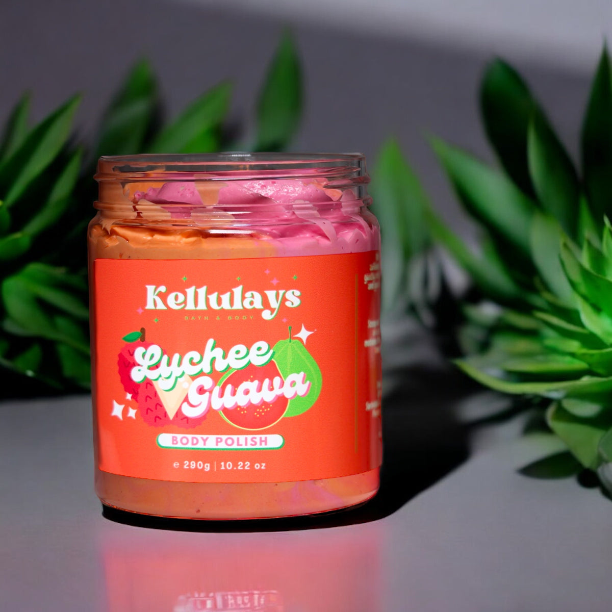Jar of Kellulays Lychee Guava Body Polish with pink and orange swirls inside, set on a reflective surface with green succulents in the background, highlighting a vibrant tropical theme.