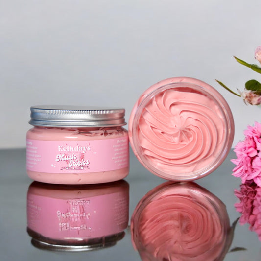 Open jar of Kellulays Musk Sticks Body Butter with a pink swirled texture, set on a reflective surface next to pink flowers, showcasing its creamy and luxurious appearance.