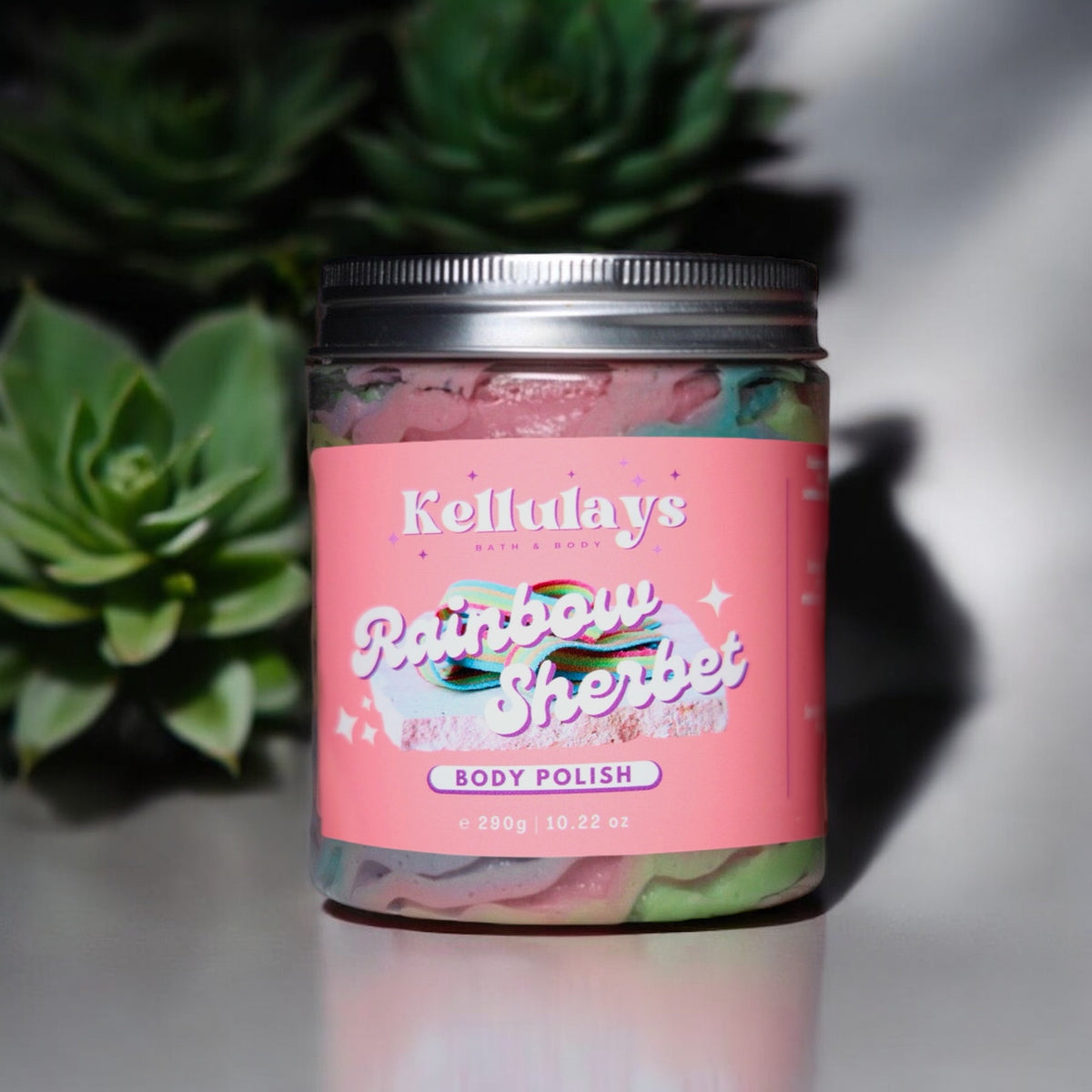 Jar of Kellulays ‘Rainbow Sherbet’ body polish with a pink label, featuring colorful swirls inside. Succulents in the background.