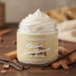 A jar of Kellulays Vanilla Sandalwood Whipped Body Butter sitting on a wooden surface, filled with a thick, creamy white swirl. Te beige label features the Kellulays logo and product name. Surrounding the jar are vanilla bean pods and sandalwood pieces, with soft steam rising in the background for a warm, cosy, dessert-like atmosphere.