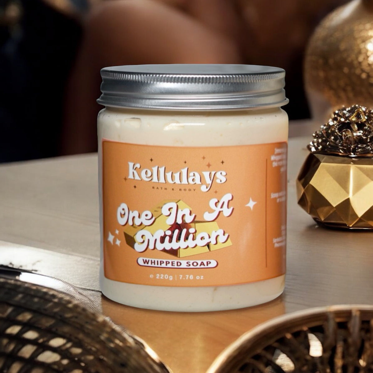 A jar of Kellulays One In A Million Whipped Soap with an orange label displaying the product name in elegant white script. The jar, filled with creamy soap, has a silver lid and is set on a wooden surface surrounded by decorative gold elements, creating a luxurious and sophisticated ambiance."
