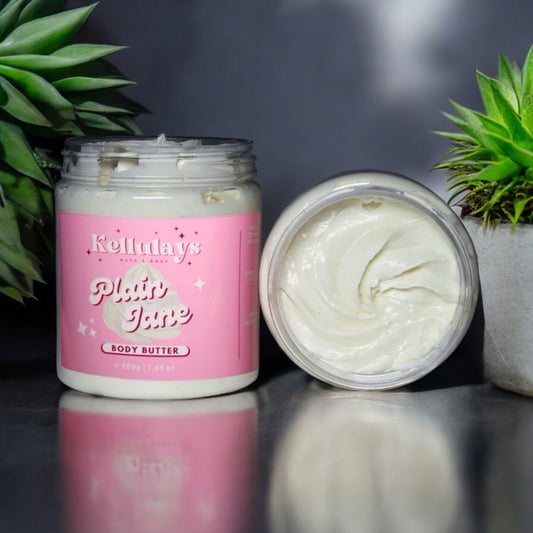 A jar of Kellulays Plain Jane Body Butter with a pink label, featuring the product name in white script. The body butter has a creamy, smooth texture and is unscented. The open jar is displayed alongside a closed jar, surrounded by small green succulent plants, against a dark, reflective surface.