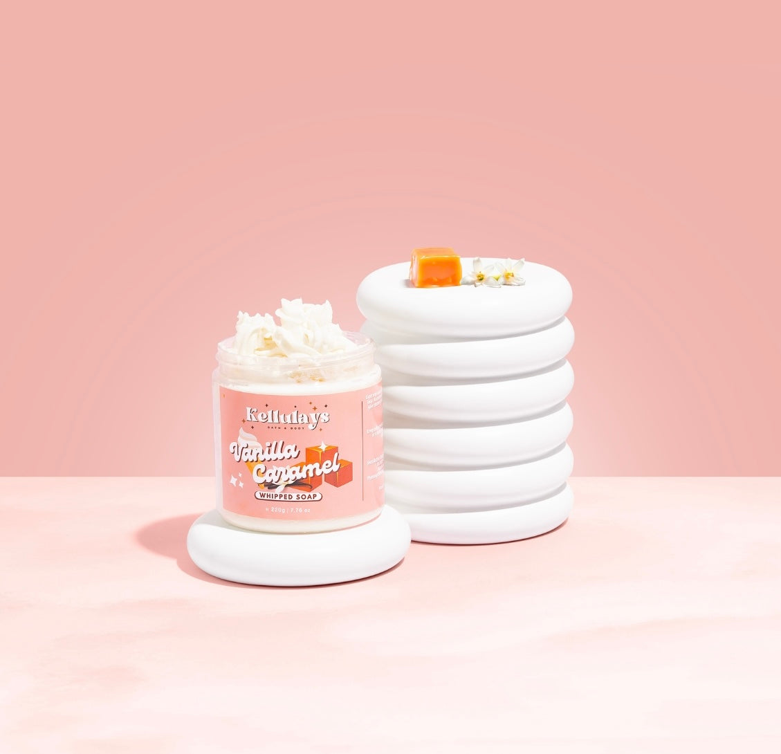 Kellulays Bath & Body Vanilla Caramel Whipped Soap. This luxurious 220g jar is styled against a blush pink background, showcasing its creamy texture. The jar features a charming label adorned with vanilla pods and caramel cubes, reflecting the indulgent scent. Beside the jar, a stack of smooth white ceramic discs holds a caramel cube and delicate white flowers, emphasizing the soap's sweet and aromatic allure.